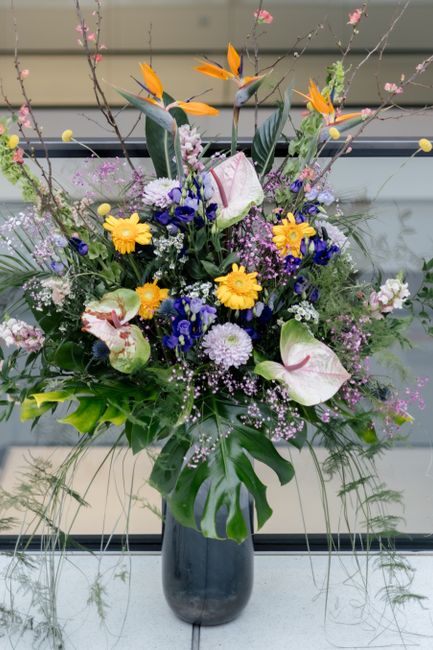 A tall, vibrant floral arrangement featuring a variety of fresh flowers in a sleek, dark vase. The bouquet includes striking orange bird of paradise flowers with elongated petals and stems, creamy whi