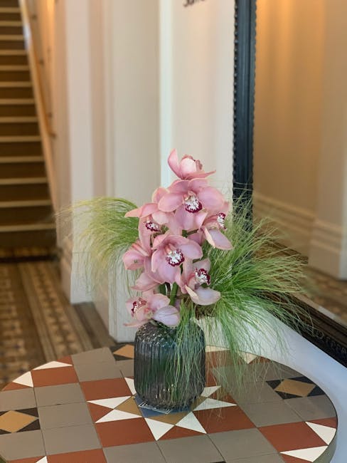 A tall, elegant floral arrangement of light pink orchids with multiple blooms cascading downward, set in a dark ribbed glass vase. The orchids are accented with wispy green grasses that add texture an